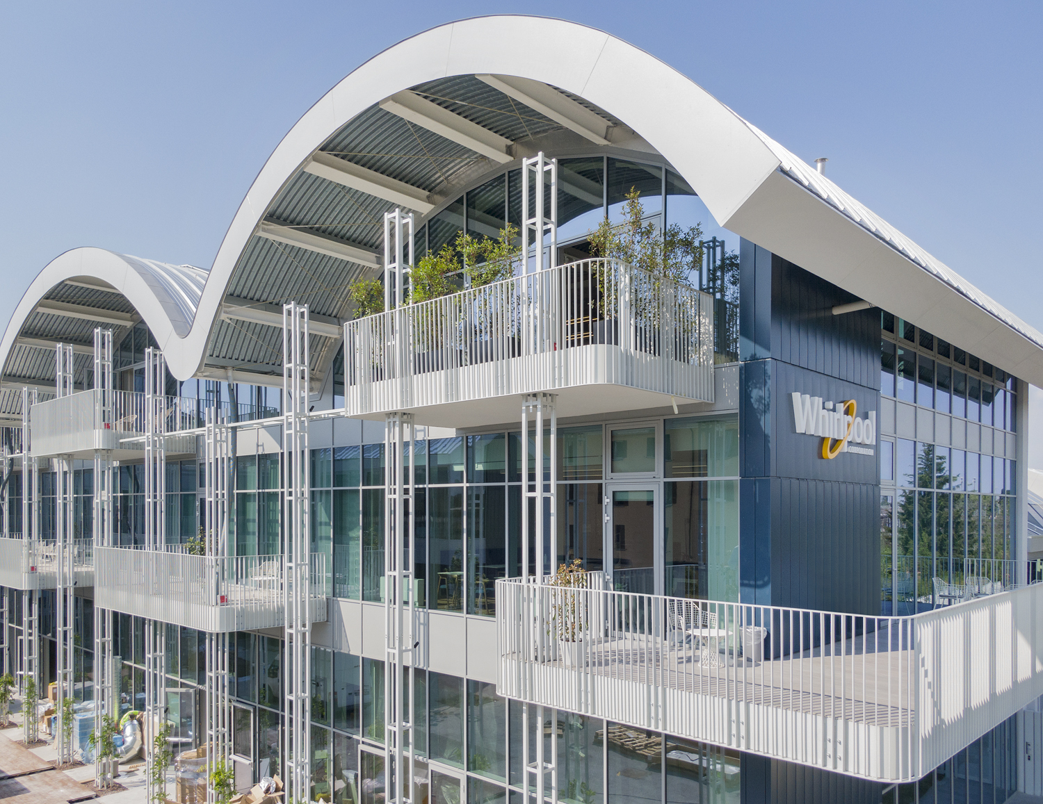 Edificio moderno multipiano con una distintiva copertura metallica a tre grandi onde simmetriche. La facciata è interamente vetrata, interrotta da balconi bianchi a sbalzo sorretti da sottili tralicci verticali in acciaio. Sul fianco scuro spicca il logo Whirlpool. Progetto Varesina 204 di carpenteria strutturale complessa realizzata da Fratelli Gelmini.