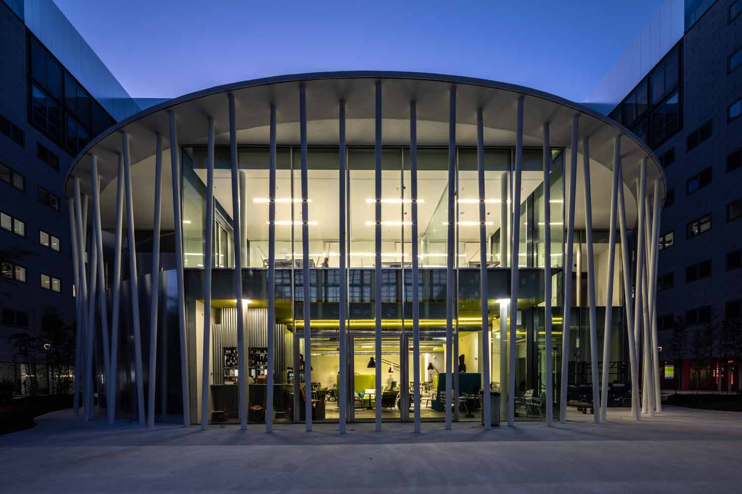 Padiglione circolare vetrato del BIM Milano Bicocca caratterizzato da una fitta serie di colonne metalliche bianche sottili e inclinate. La struttura in acciaio realizzata da Fratelli Gelmini circonda l'edificio illuminato all'imbrunire.